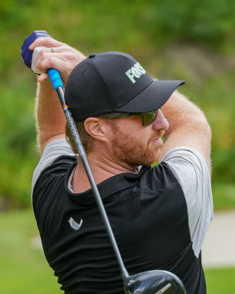 A male golfer in a black and gray shirt, black Dirbie "FORE" Golf Cap, and sunglasses swings a driver golf club on a golf course with green grass in the background.
