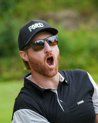 A man on a golf course wearing a black "Dirbie" FORE! Golf cap, dark sunglasses, and a black and white polo shirt, with his mouth open shouting "FORE".